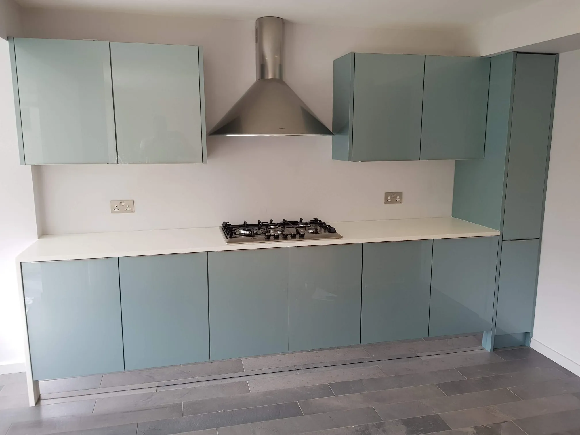 Duck egg blue gloss handleless kitchen with white worktop, gas hob and chimney hood
