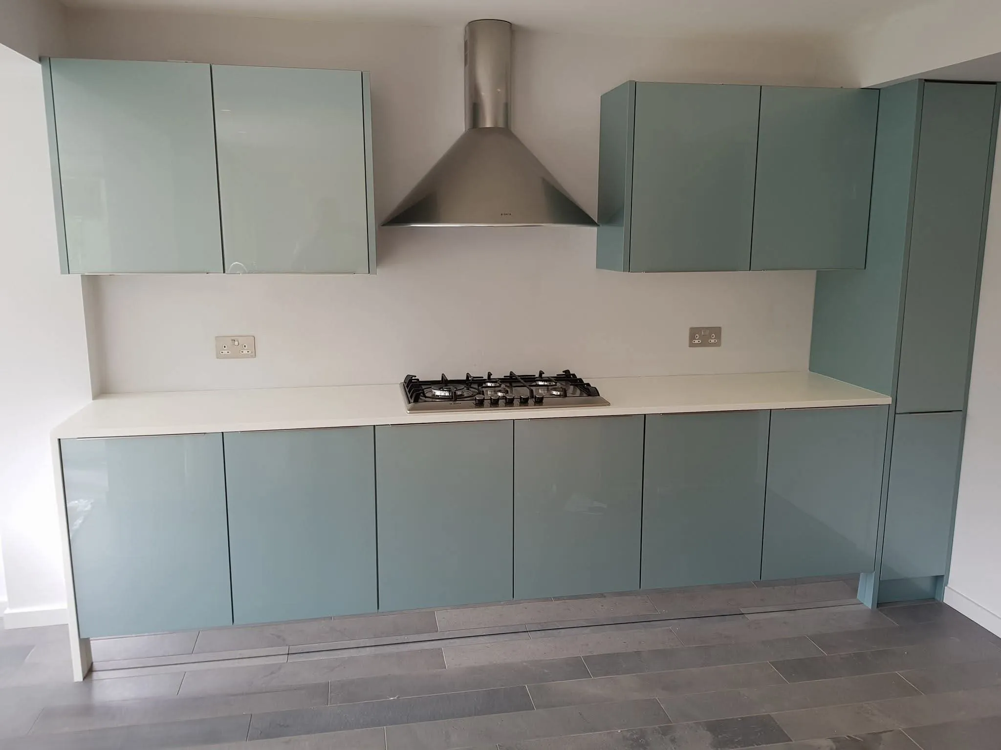 Pale teal gloss handleless kitchen with white worktop, gas hob and stainless chimney hood